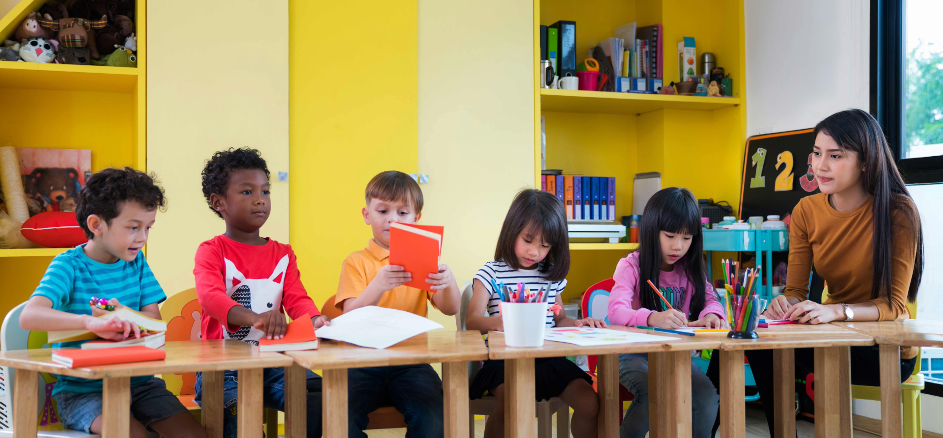 A kindergarten teacher sits at a table with five students